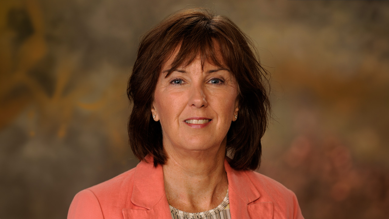 A brown-haired woman in an orange blazer poses for headshot against a brown background.