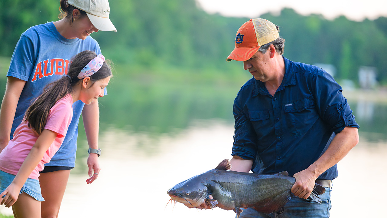 A man holds a large catfish while a girl and young woman examine it