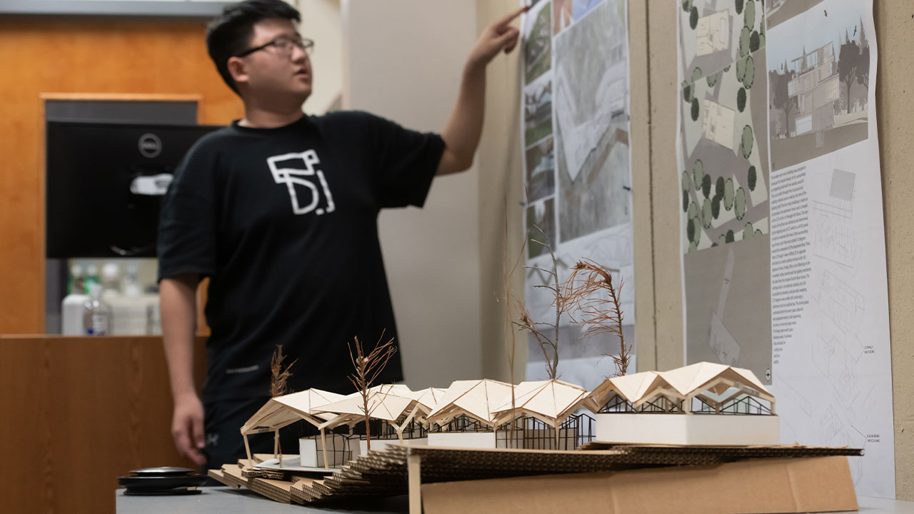 A 3D building model is at the forefront while a student works on a white board 