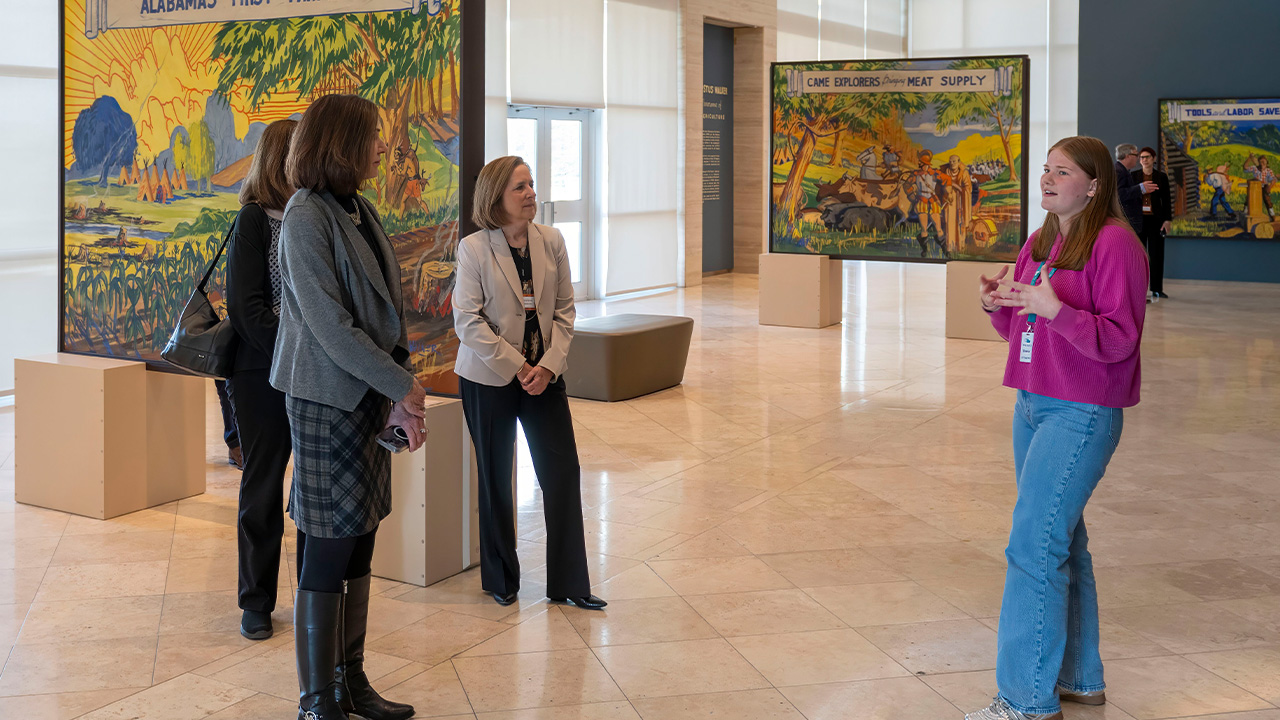 A young woman in a pink sweater gives a tour to visitors in front of art at the museum