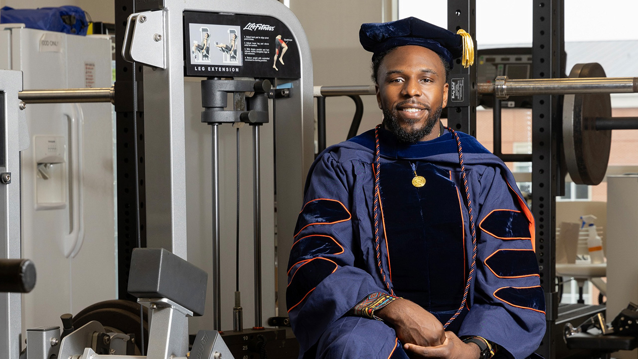 Mason sits on a weigh machine wearing a cap and gown