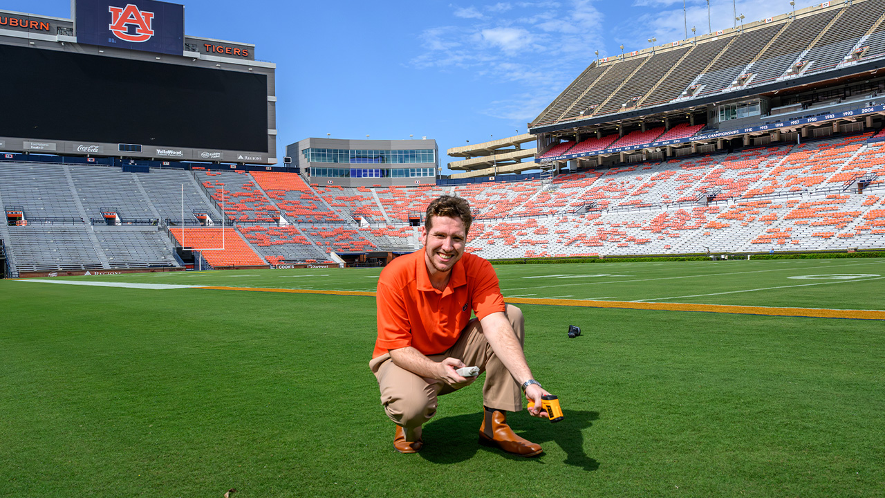 Brandon kneels in the middle of the football field in the stadium holding an instrument with a small screen