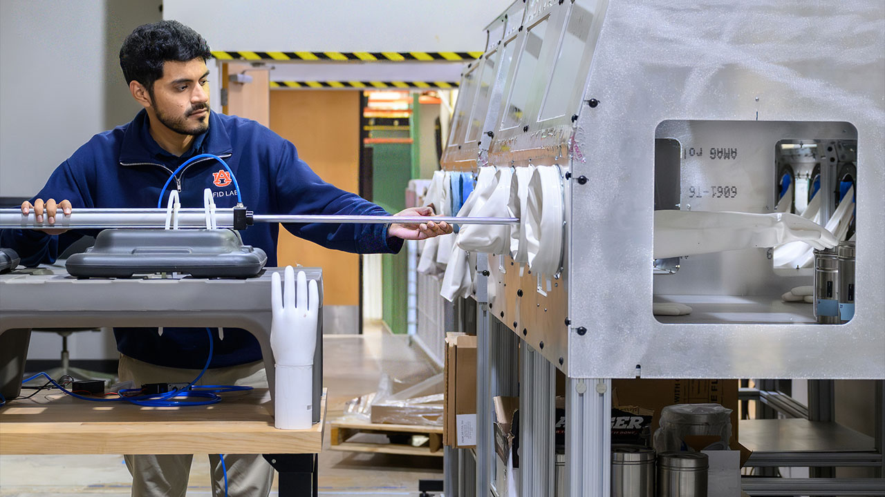 A young man operates a robotic arm, showing how it is used with a replica storage facility to test repeated handling of RFID technology.
