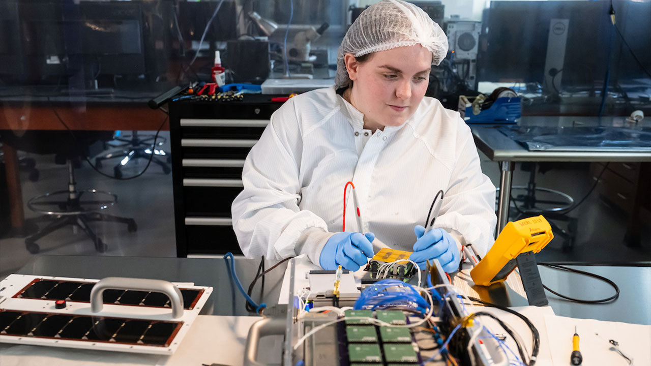 A young female student, wearing a hairnet, gloves and white coat because she is in a clean room, works on a CubeSat in the AU Small Satellite Program lab.