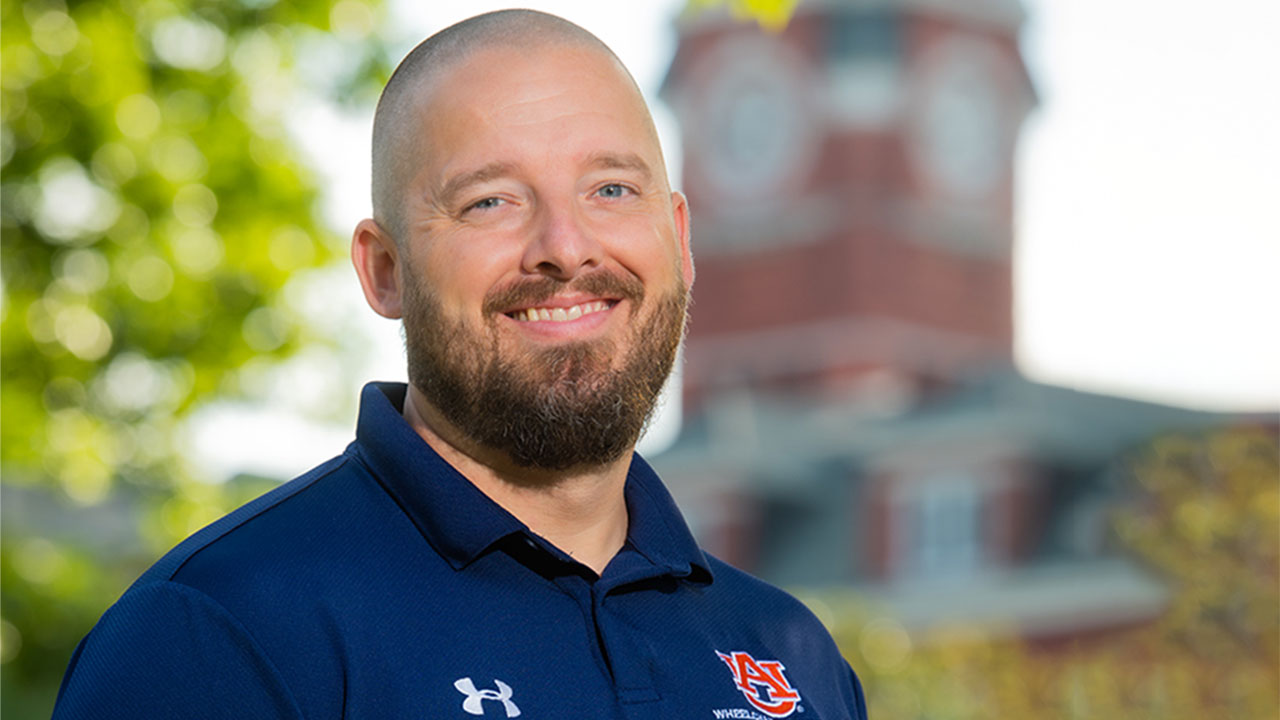 Coach Robb Taylor stands smiling in a blue Auburn polo shirt, with Samford Hall shown behind his right shoulder.