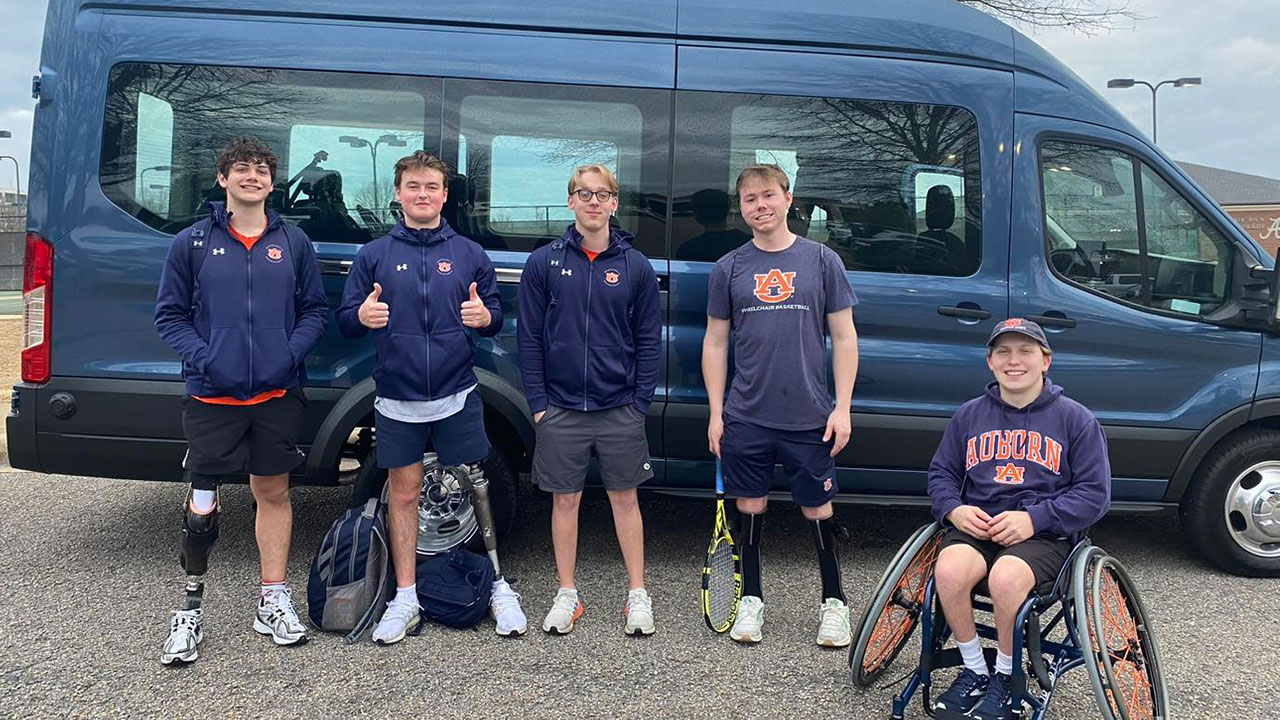 Five members of Auburn's Wheelchair Basketball Team pose in front of a blue van. Four of the young men are standing and the fifth man, who is pictured on the far right, is seated in a wheelchair.