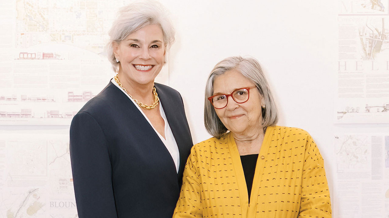 Two women, Beth Stukes and Cheryl Morgan, stand side by side smiling in front of an architecture-themed backdrop.