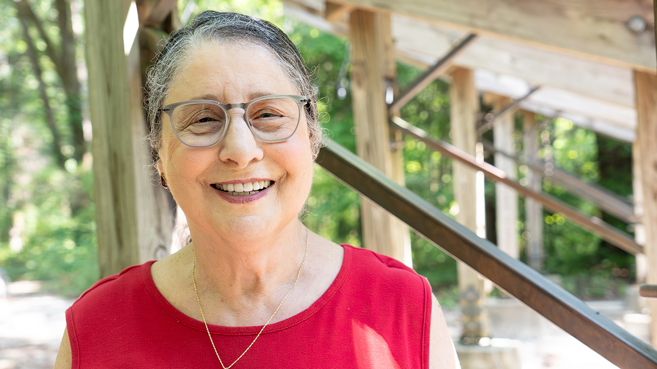 Emily Kling, smiling and wearing a red t-shirt, stands near the amphitheatre in the Kreher Preserve and Nature Center with trees dotting the background. 