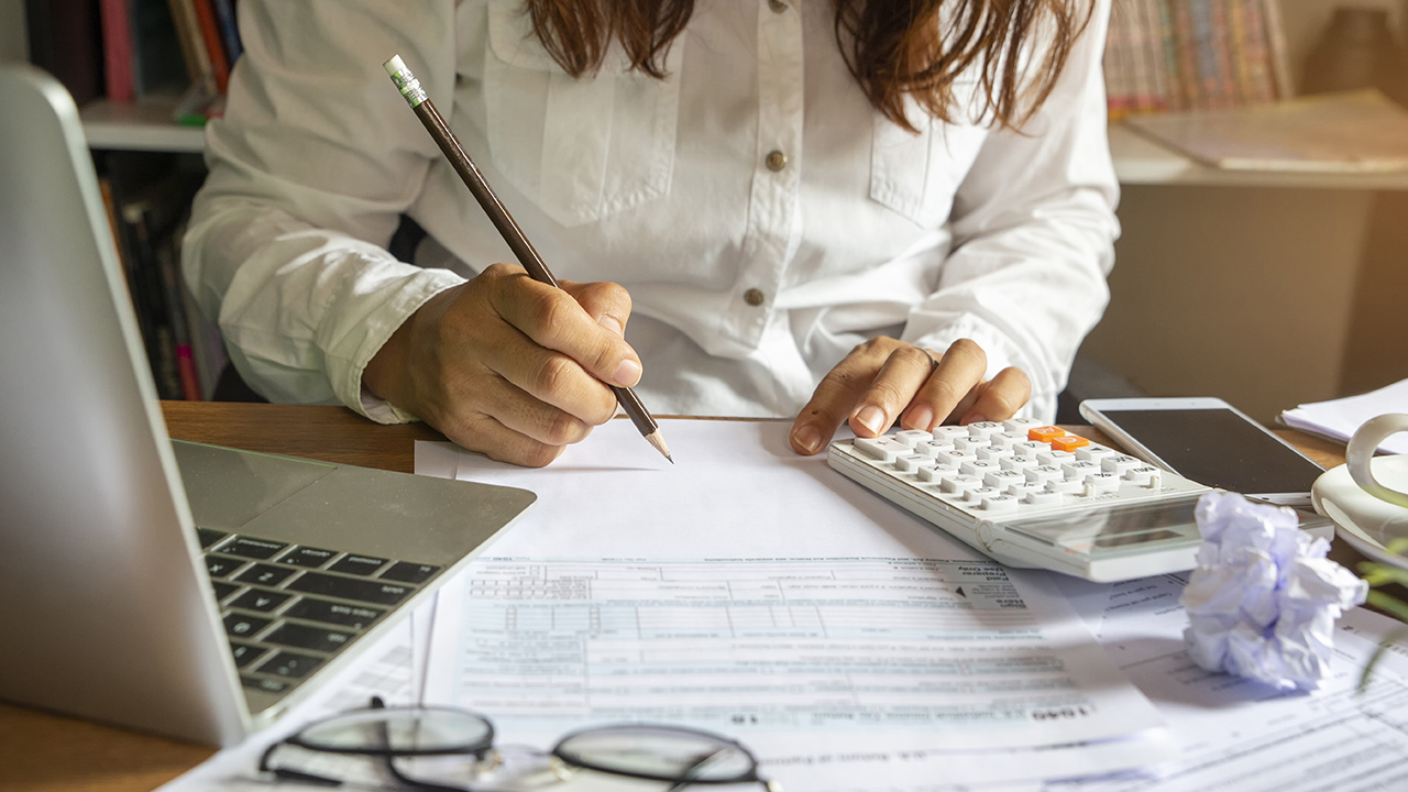 A woman at a desk writes on a piece of paper. A calculator, tax forms, and eyeglasses can be seen in the foreground.