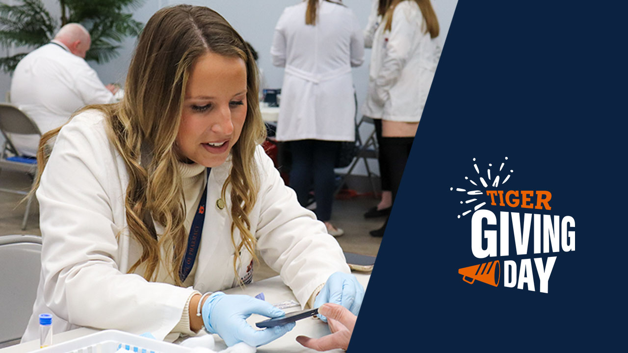 Pharmacy student in a white coat sits in front of an unseen patient and holds a glucose monitor.