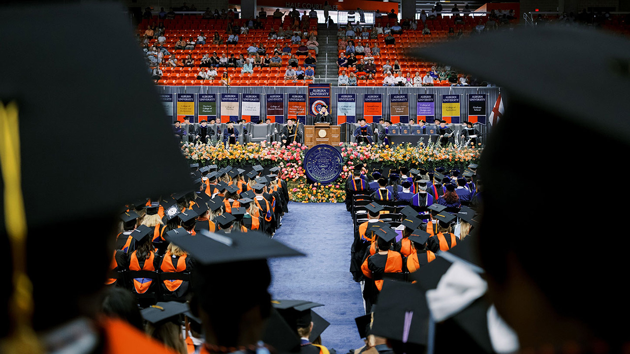 A wide angle shot with the backs of Auburn graduates in the foreground and the commencement speaker podium in the background
